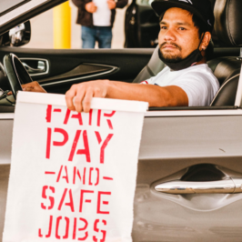 A man of color holding a sign that reads "FAIR PAY AND SAFE JOBS" while sitting in the driver's seat of a car, with a focused expression on their face. Advocates in Denver rally at the airport. photo credit: Gig Workers Rising
