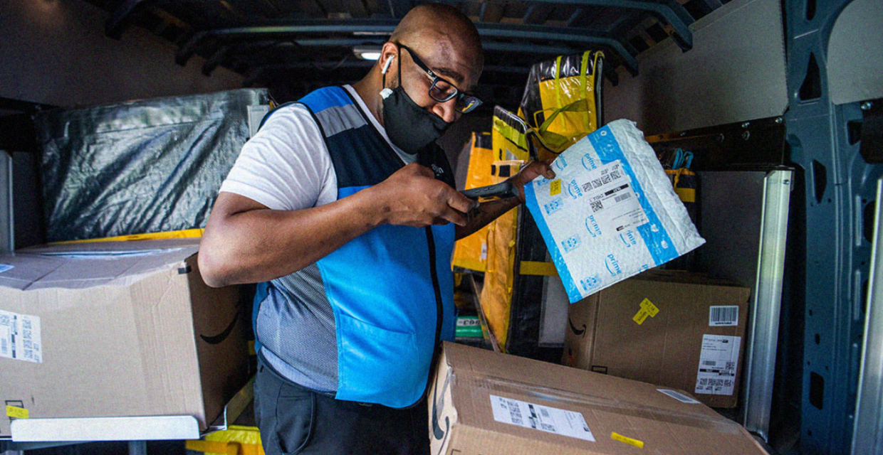 An older African American man scans packages in the back of a van. He wears a blue and grey Amazon vest.