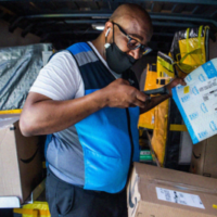 An older African American man scans packages in the back of a van. He wears a blue and grey Amazon vest.