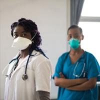 Two black women in a hospital wearing masks. Woman on left is in a white lab coat and woman on right is in blue scrubs. Both facing camera.