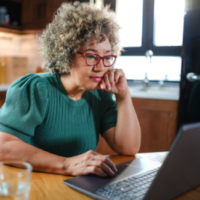 A concerned mature woman with curly gray hair reviews paperwork at her kitchen table. She holds a document while looking at it intently, surrounded by a laptop, calculator, and papers.