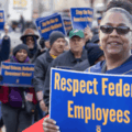 A smiling Black woman wears an AFGE jersey and holds a sign that reads, 'Respect Federal Employees' in front of a crowd of marchers. Photo credit: American Federation of Government Employees, AFL-CIO