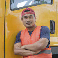 A truck driver wearing an orange safety vest poses in front of a yellow big rig truck.