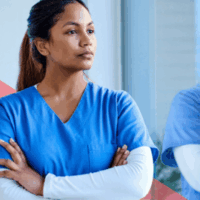 A medical worker wearing blue scrubs, looks thoughtfully out of a window