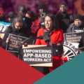 A woman among a crowd, speaks at a podium bearing a sign that reads "EMPOWERING APP-BASED WORKERS ACT".