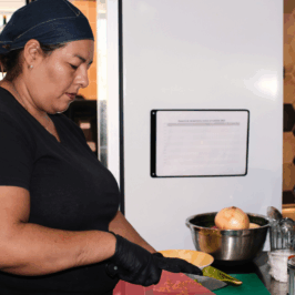 A middle-aged Latina woman chops jalapenos in a restaurant kitchen. She wears a denim bonnet, black t-shirt, and black latex gloves.