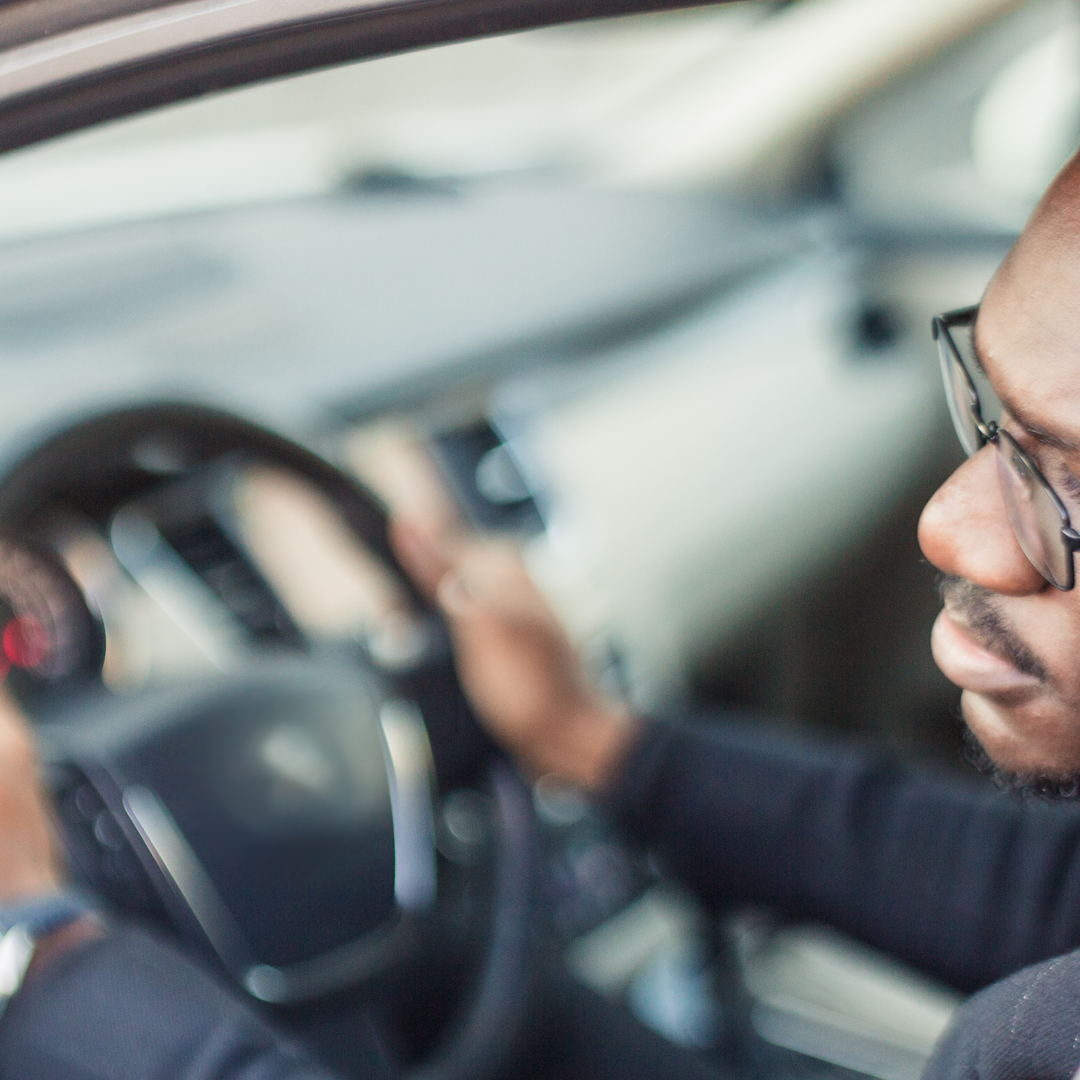 A Black man in a car looks out to his left-side mirror.